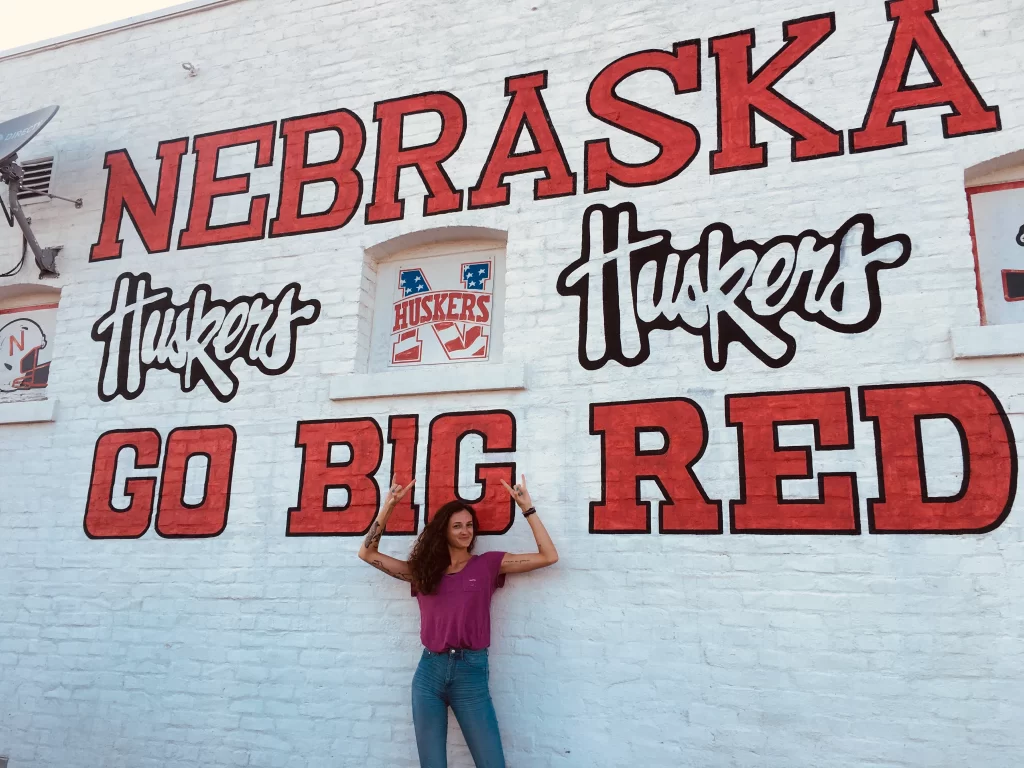 Woman with her arms up against a white wall with red writing that says "Nebraska Huskers, go big red"